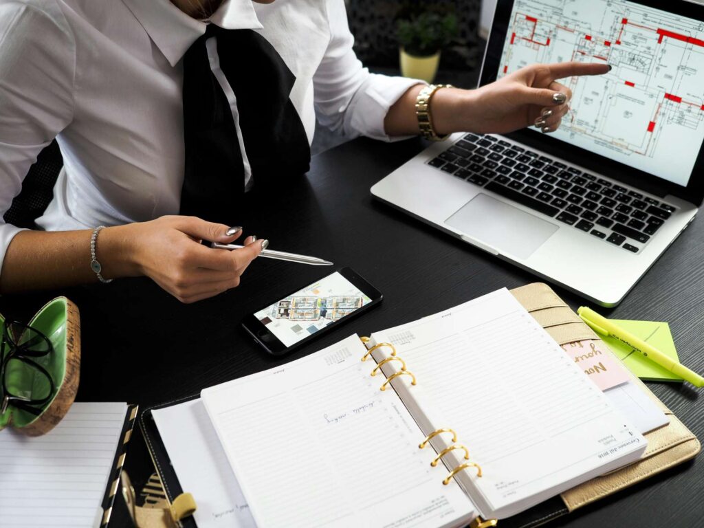 A professional woman at a desk pointing to architectural plans displayed on a laptop screen. She holds a pen in one hand and has a smartphone on the desk showing a map or floor plan. An open planner, notepad, highlighter, and eyeglasses are also on the desk, indicating a workspace for planning or consulting.