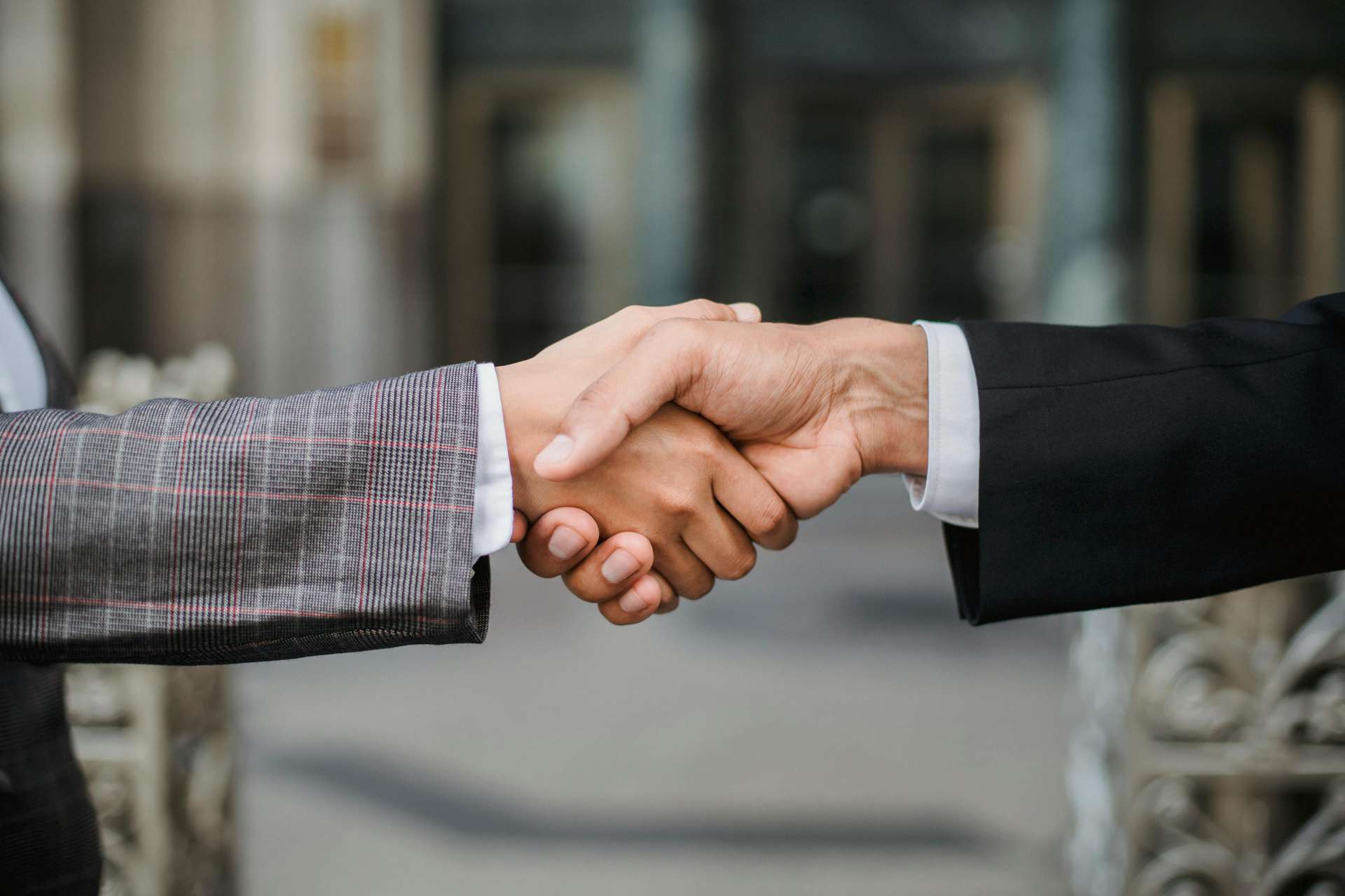 A close-up of two individuals shaking hands, symbolizing agreement or partnership. One person wears a suit with a plaid pattern, and the other wears a plain black suit. The background is blurred, suggesting an outdoor or professional setting.